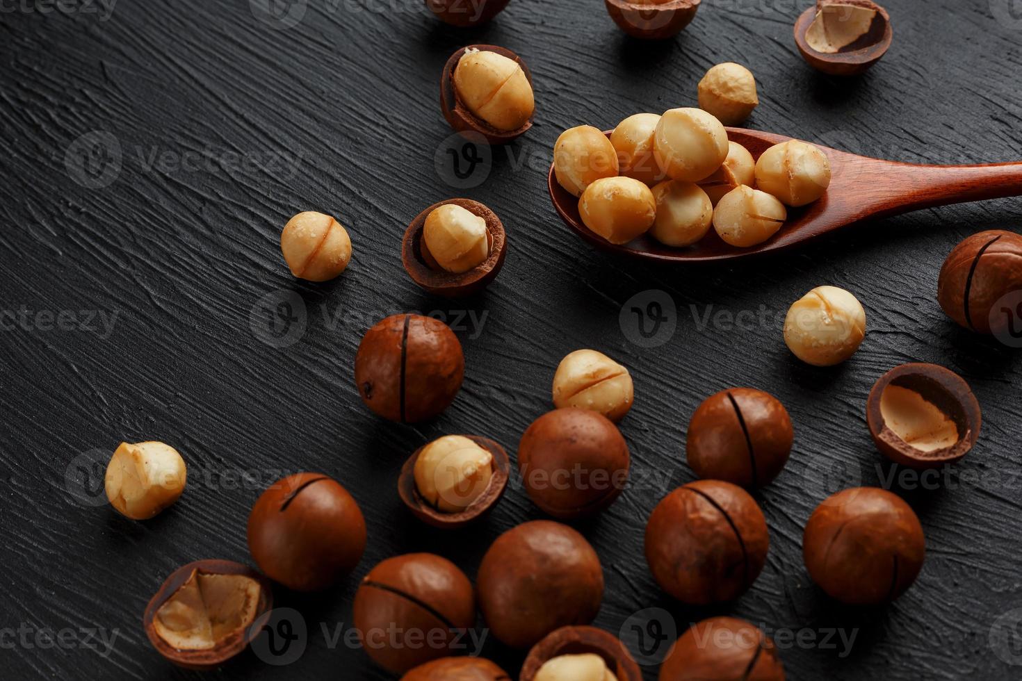 sgusciate macadamia Noce e pelato macadamia Noce su un' nero strutturale sfondo nel un' di legno cucchiaio foto