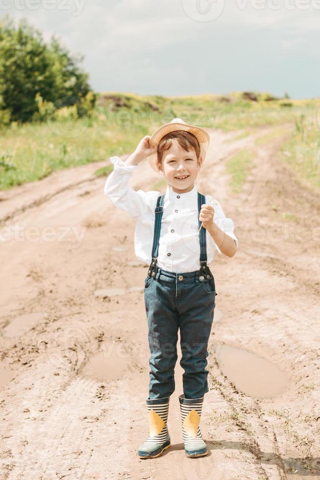 poco contadino su un' estate campo, carino poco ragazzo nel un' cannuccia cappello. ragazzo nel un' cappello con un' fiore sta nel un' campo. ritratto di un' villaggio ragazzo foto