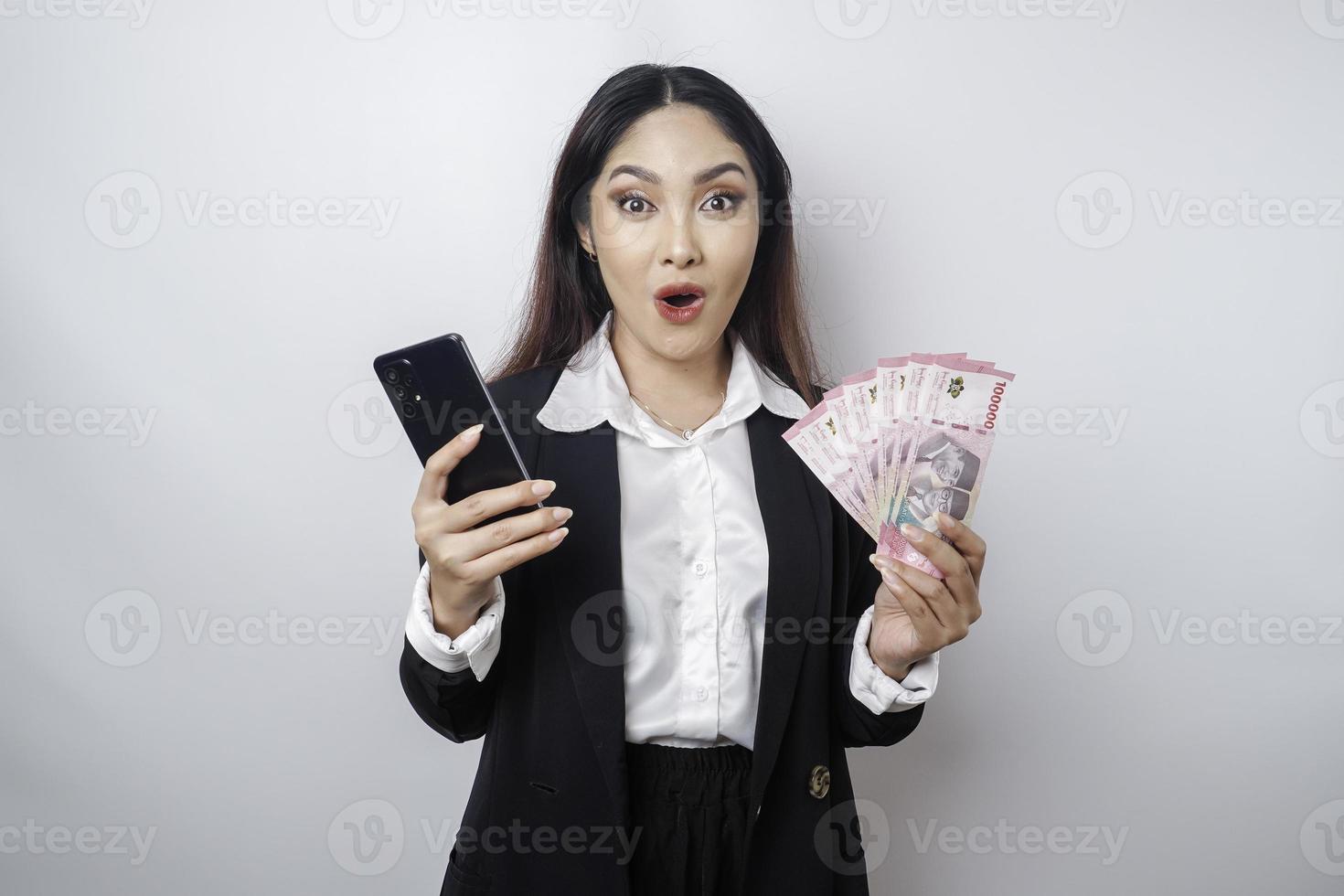 sorpreso asiatico donna d'affari indossare nero completo da uomo Tenere sua smartphone e i soldi nel indonesiano rupia, isolato di bianca sfondo foto