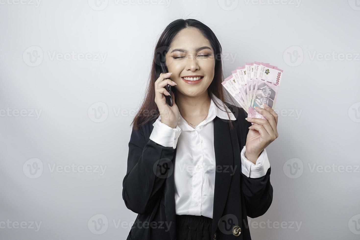 un' contento giovane donna d'affari è indossare nero completo da uomo, Tenere sua Telefono e i soldi nel indonesiano rupia isolato di bianca sfondo foto