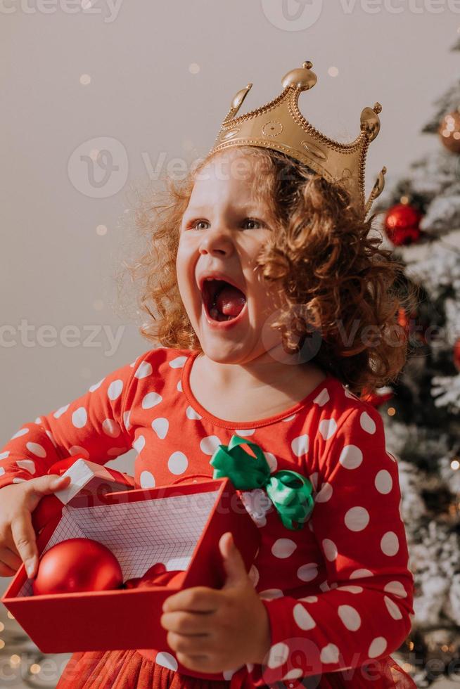 poco dai capelli ricci ragazza nel un' carnevale vestito nascosto sua viso dietro a brillante Natale albero giocattoli stelle. bambino nel un' rosso vestito con un' Santa Stampa su il sfondo di un' Natale albero. alto qualità foto