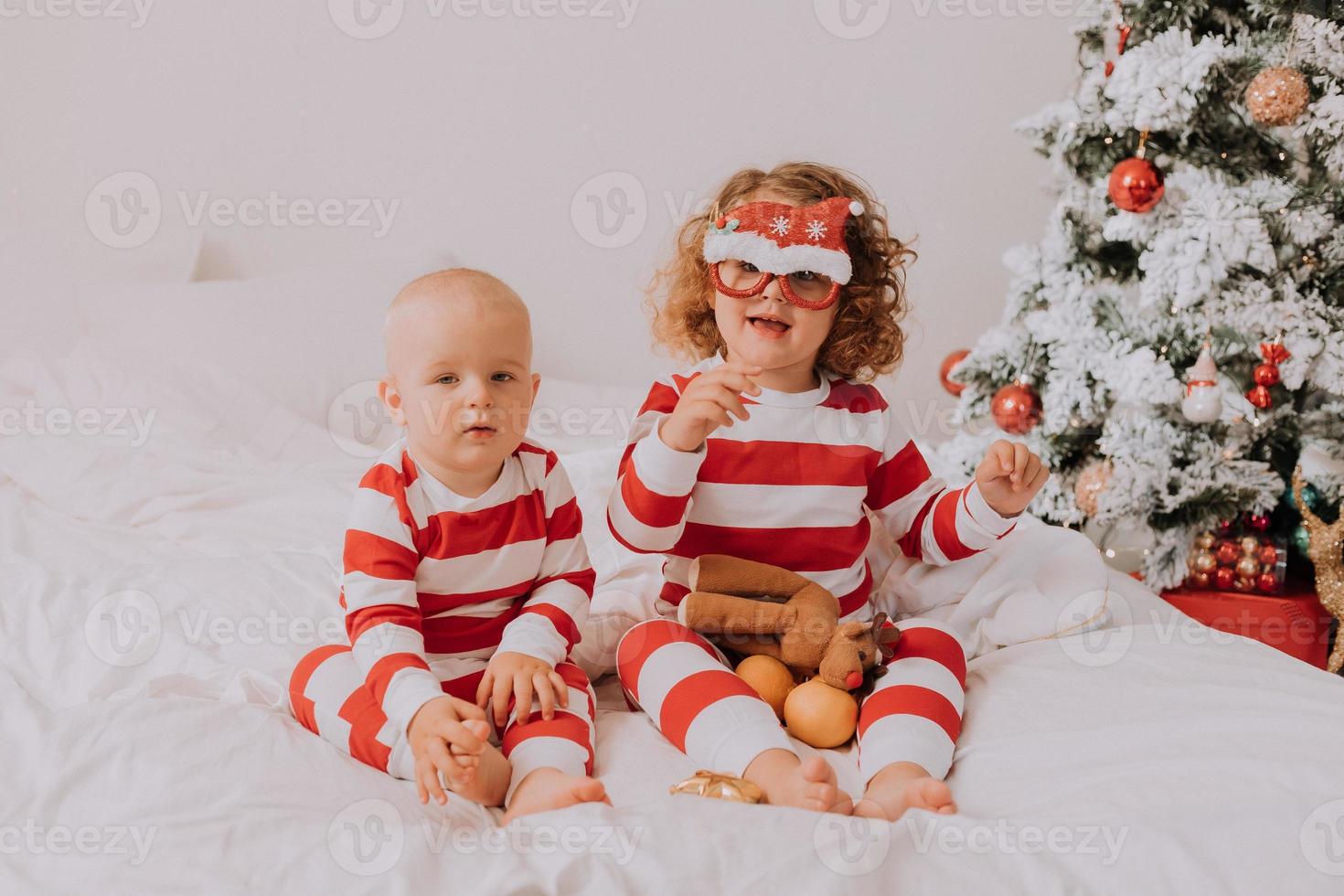 bambini nel rosso e bianca pigiama provare su divertente bicchieri con Santa Claus seduta nel letto. stile di vita. fratello e sorella festeggiare Natale. ragazzo e ragazza siamo giocando a casa. alto qualità foto