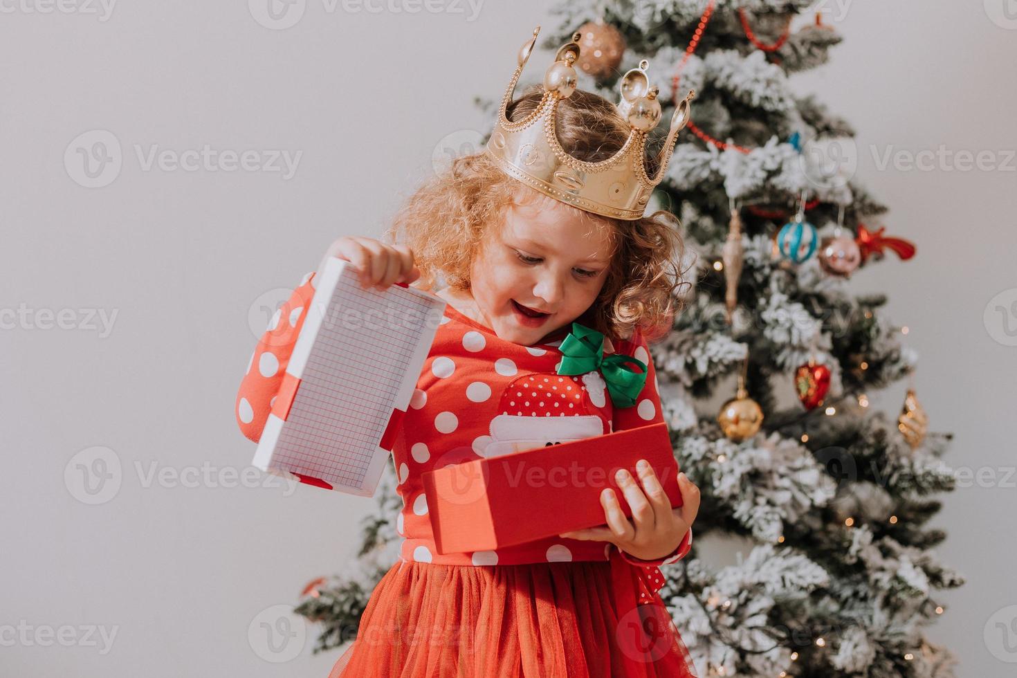 poco dai capelli ricci ragazza nel un' carnevale vestito nascosto sua viso dietro a brillante Natale albero giocattoli stelle. bambino nel un' rosso vestito con un' Santa Stampa su il sfondo di un' Natale albero. alto qualità foto