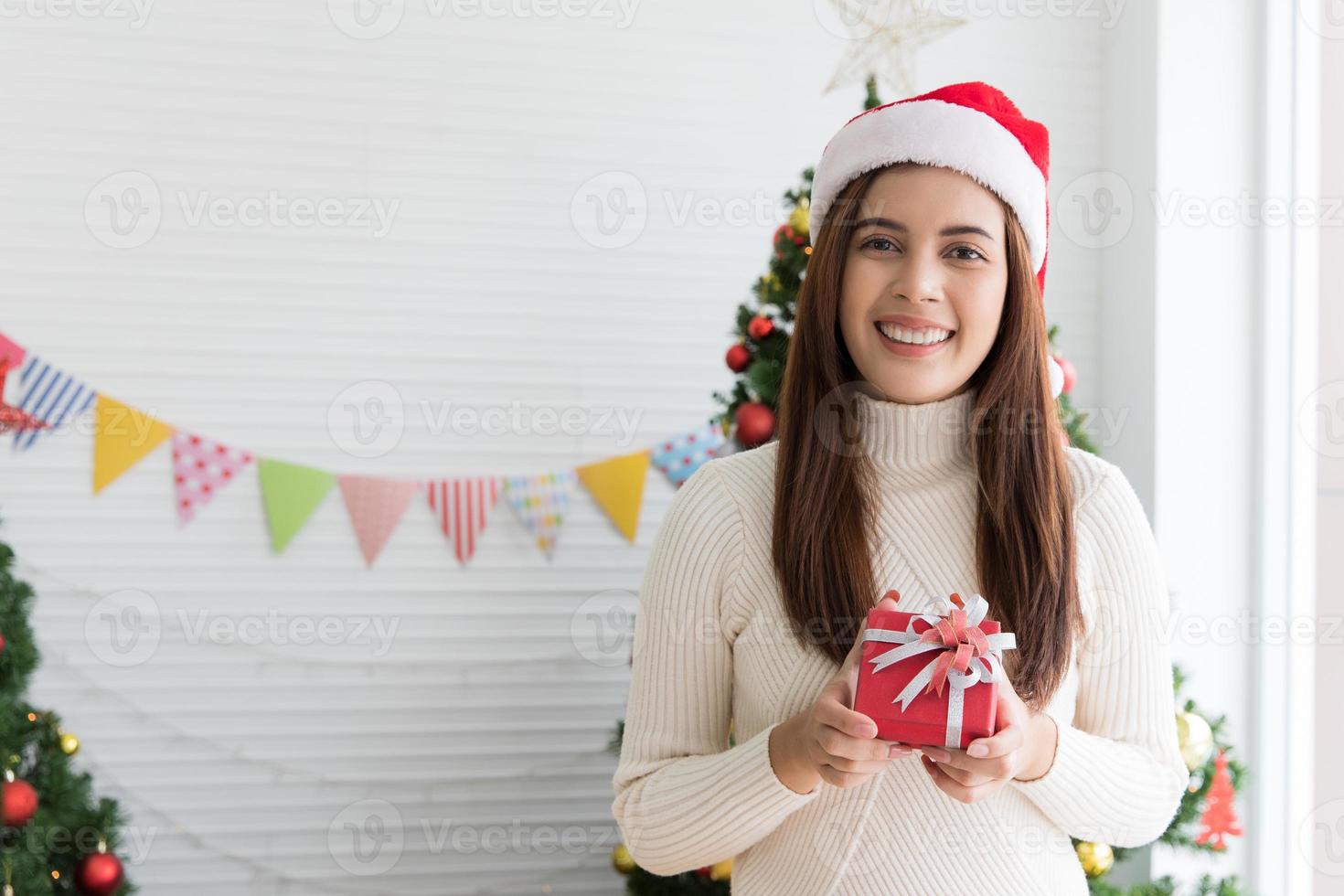 sorridente brunetta donna nel maglione e Santa cappello Tenere piccolo regalo scatola e gioisce al di sopra di Natale decorativo bianca sfondo foto