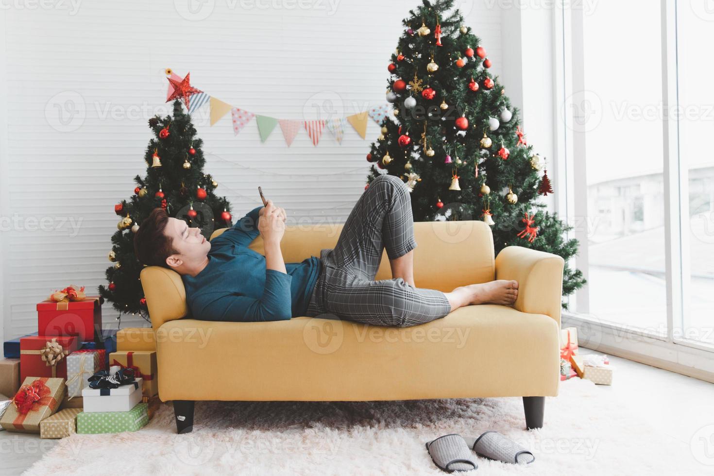 un' singolo uomo sembra solitario utilizzando il suo Telefono e posare giù su divano nel vivente camera decorato con Natale alberi e regalo scatole foto