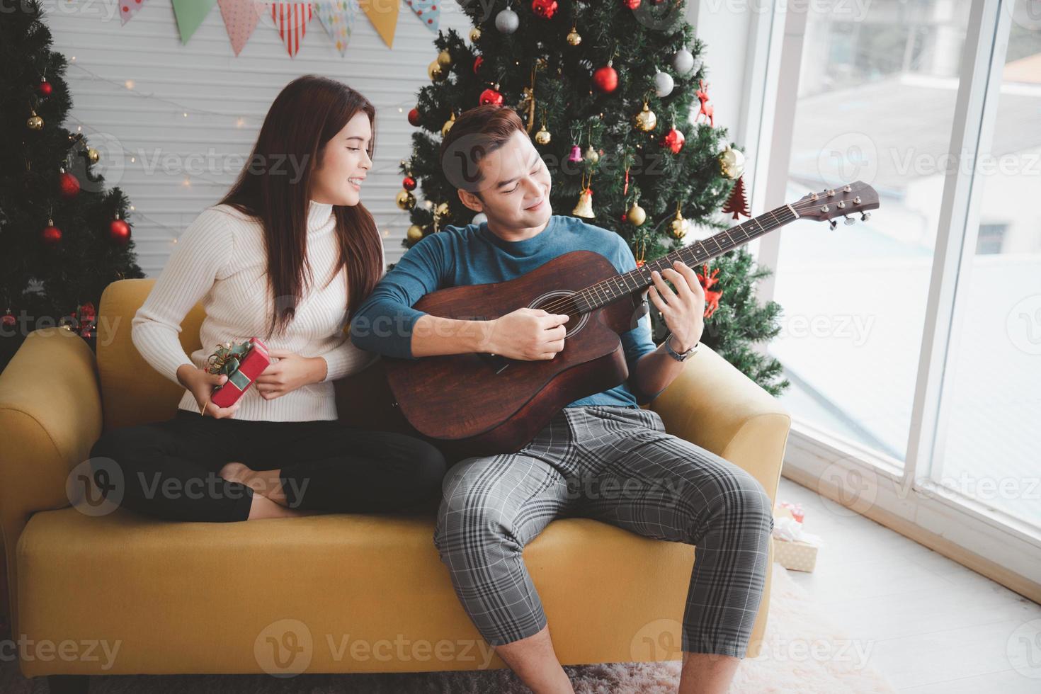 un' uomo giocando chitarra per donna amante durante festeggiando Natale vacanza insieme nel vivente camera con Natale albero ornamento decorazione foto