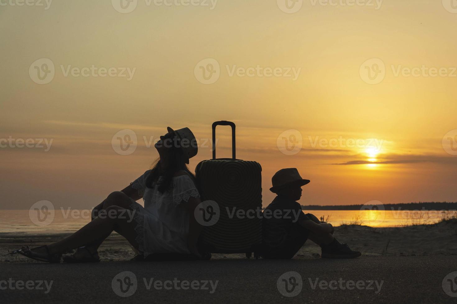 madre e giovane figlio con un' valigia su il sfondo di il tramonto in attesa .un viaggio per il mare, un' vacanza, il oceano, un' viaggio per caldo paesi foto