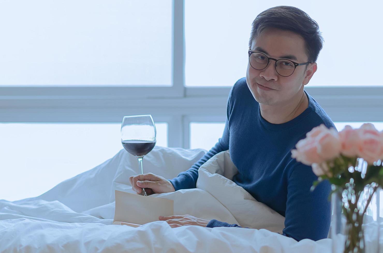 un' sorridente persona quello lettura libro su letto con un' bicchiere di rosso vino. bene mentale Salute concetto. foto