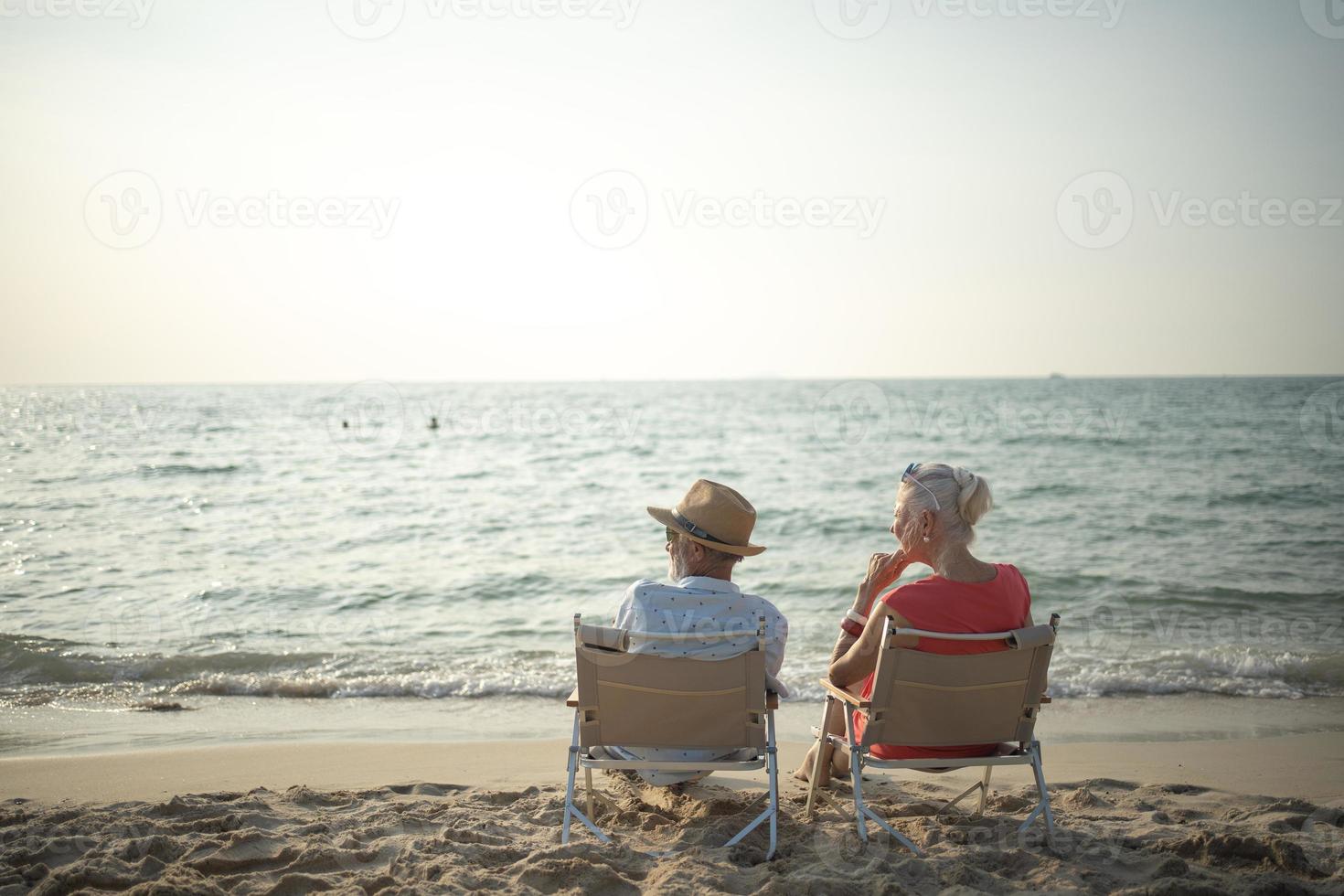 Due anziano uomini e donne sedersi sedia a il spiaggia parlando e Guardando il sole e il mare su loro estate vacanza e essi Sorridi e godere loro vacanza. foto