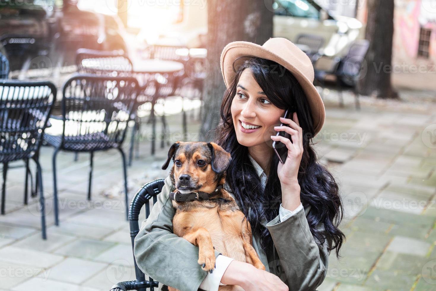 attraente giovane donna seduta nel caffè negozio, rilassante e Tenere sua cane mentre parlando su il Telefono. animale domestico amichevole caffetteria, bellissimo ragazza con sua cane ubicazione a un' caffè negozio e bevanda caffè. foto