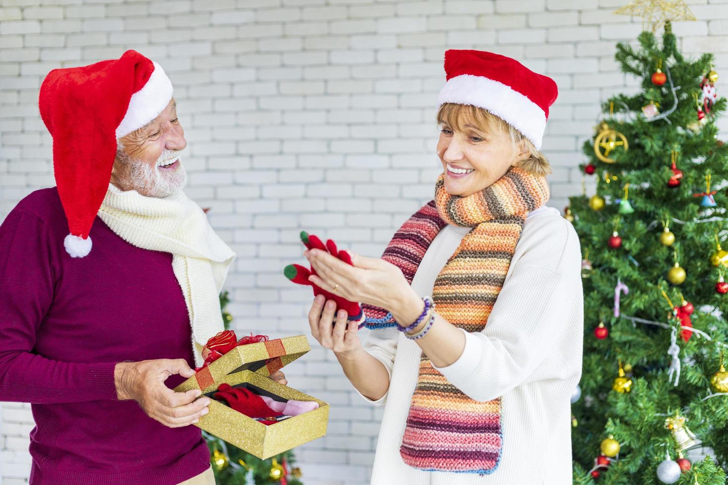 contento sorridente anziano caucasico coppia festeggiare Natale insieme mentre scambiare loro presente nel felicità e eccitazione a casa con rosso Santa cappello e Natale albero foto