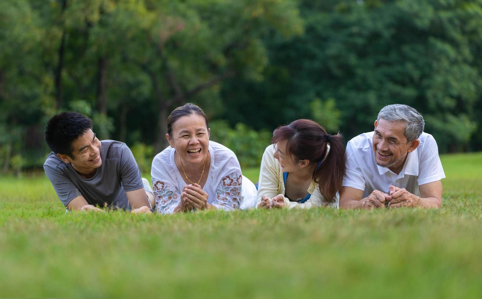 gruppo di asiatico famiglia con padre, madre, figlio e figlia dire bugie giù insieme su il erba prato e ridendo a il pubblico parco durante fine settimana attività per bene mentale Salute e ricreazione concetto foto