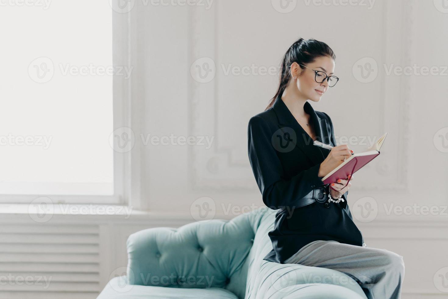 attraente donna d'affari di successo in abiti eleganti registra le informazioni nel blocco note, si siede sul retro del divano, posa nell'ufficio a casa, pianifica il processo di lavoro, fa una lista da fare, annota i punti foto