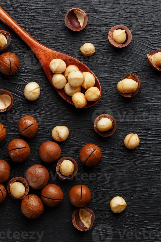 sgusciate macadamia Noce e pelato macadamia Noce su un' nero strutturale sfondo nel un' di legno cucchiaio foto