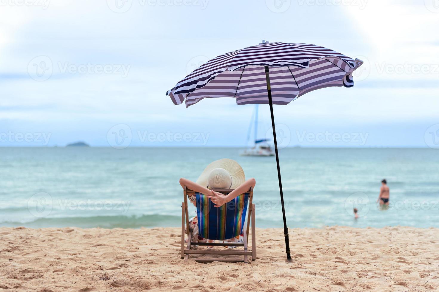 bellissimo giovane asiatico donna con cappello braccio su rilassante su spiaggia sedia nel il sole ombra ombrello, estate contento vacanza concetto. foto