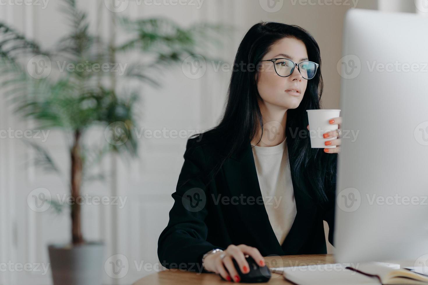 occupata giovane donna amministrativa pone al desktop, beve caffè caldo dalla tazza di carta, vestita in modo formale, sviluppa strategia aziendale, pianifica l'avvio, coinvolta nel processo di lavoro. concetto di occupazione foto