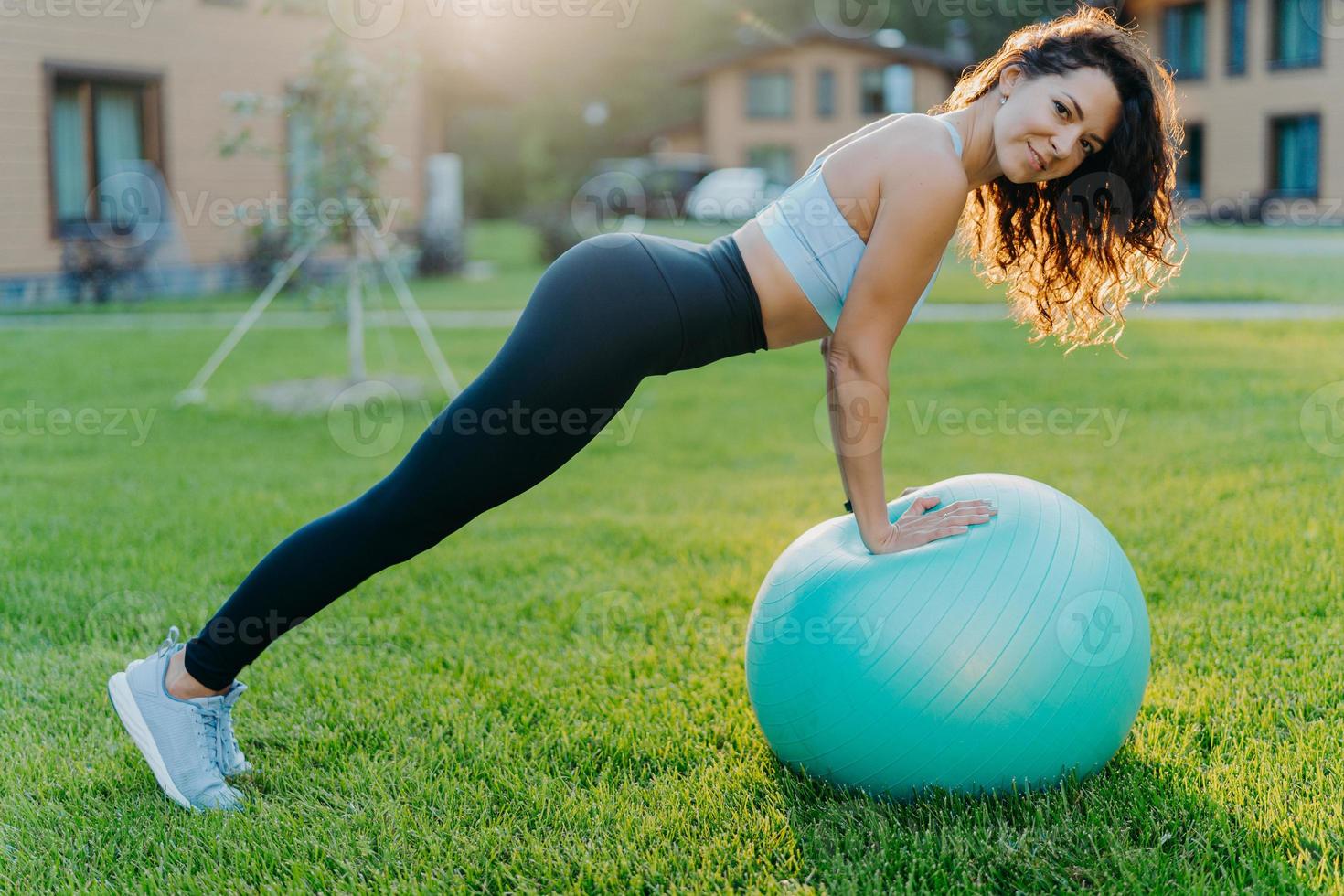 il colpo orizzontale di una donna piuttosto magra fa esercizi fisici con la palla fitness ha una figura perfetta indossa abiti sportivi pone sul prato verde all'aperto gode di un allenamento fisico. pilates e concetto di yoga foto