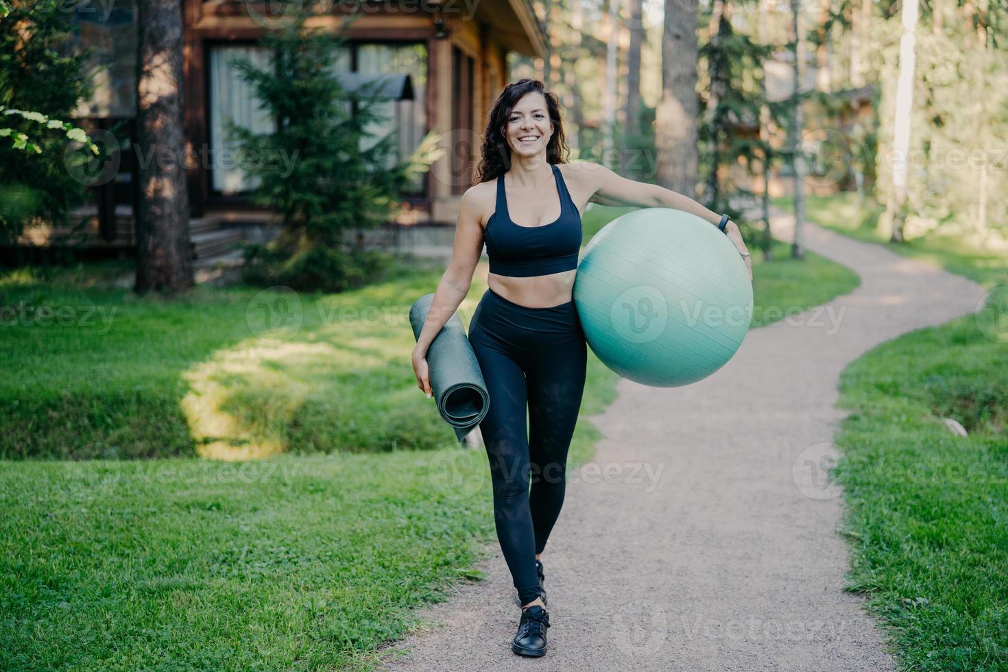 colpo all'aperto di felice donna bruna sportiva cammina sul sentiero vicino alla foresta, tiene karemat arrotolato e palla fitness, vestito con top corto, leggings e scarpe da ginnastica, ha allenamenti regolari all'aria aperta foto