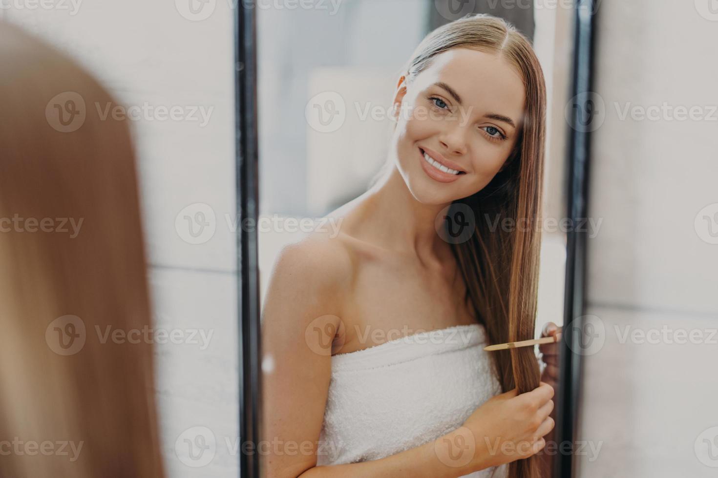 felice e bella giovane donna guarda il suo riflesso nello specchio, ha i capelli lunghi ben curati, usa la spazzola per capelli, avvolta in un asciugamano da bagno, fa l'acconciatura. concetto di donne, bellezza, acconciatura e cura di sé foto