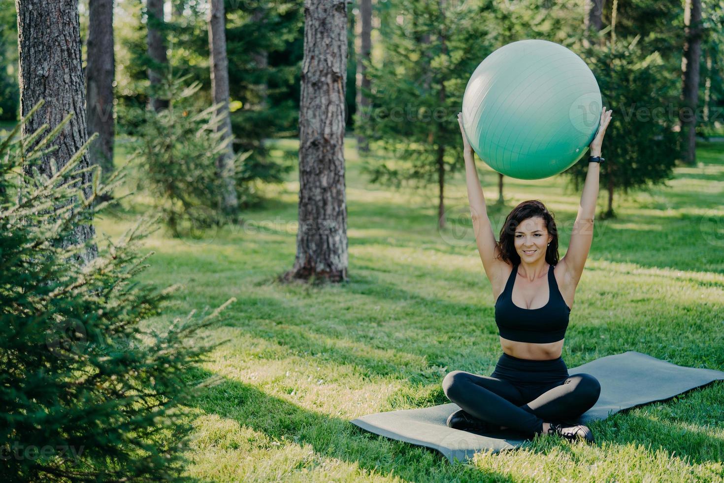 donna snella bruna solleva la palla fitness sopra la testa, si siede nella posa del loto su karemat, vestita con abiti attivi, pratica yoga all'aperto, respira aria fresca nella foresta. fit lady esercizi con palla da ginnastica foto