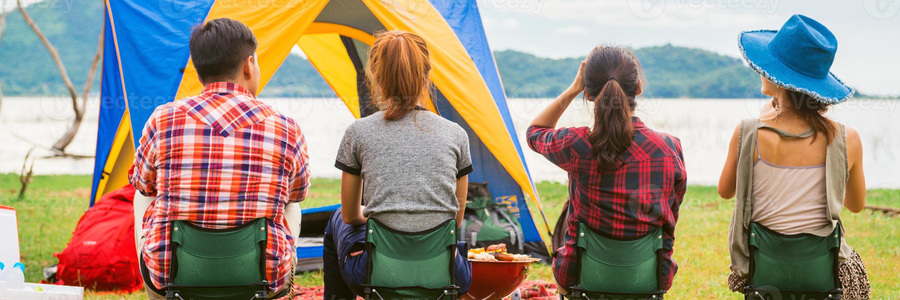 gruppo di uomo e donna godere campeggio picnic e barbecue a lago con tende nel sfondo. giovane misto gara asiatico donna e uomo. panoramico striscione. foto