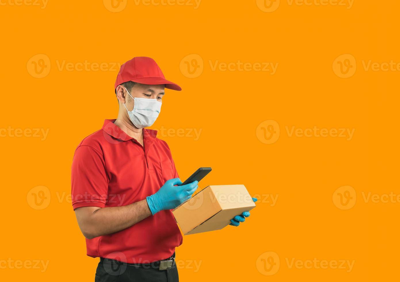 asiatico consegna uomo lavoratore indossare chirurgico maschera, medico guanti nel rosso uniforme isolato su giallo sfondo, utilizzando mobile controllo azione e hold pacco scatole per invio o veicolare pacchi di posta foto