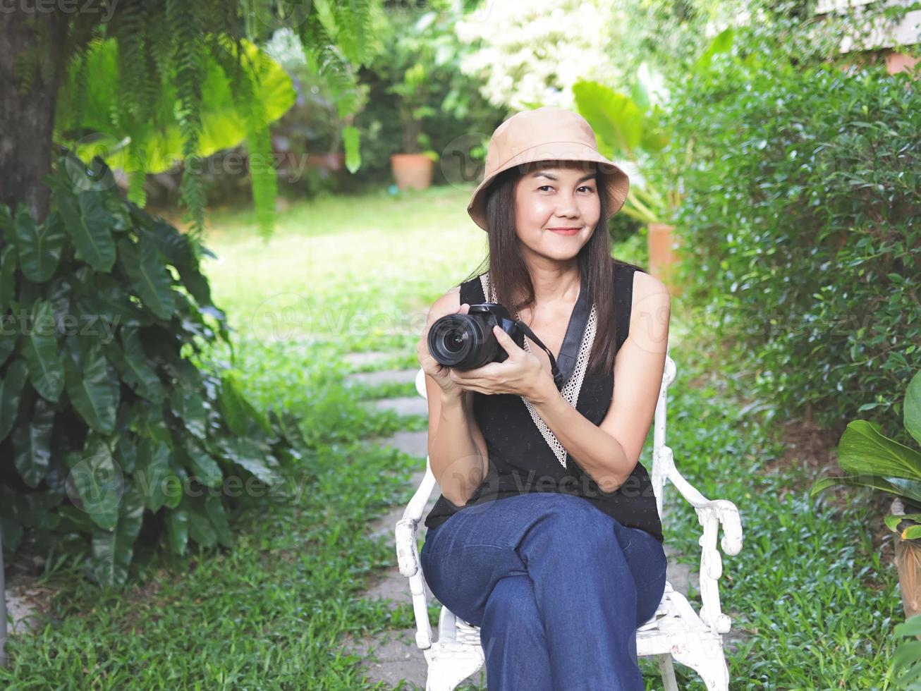 asiatico donna, indossare cappello ,Nero superiore senza maniche e blu denim pantaloni, seduta su bianca sedia nel il giardino e Tenere dslr telecamera, sorridente felicemente. foto