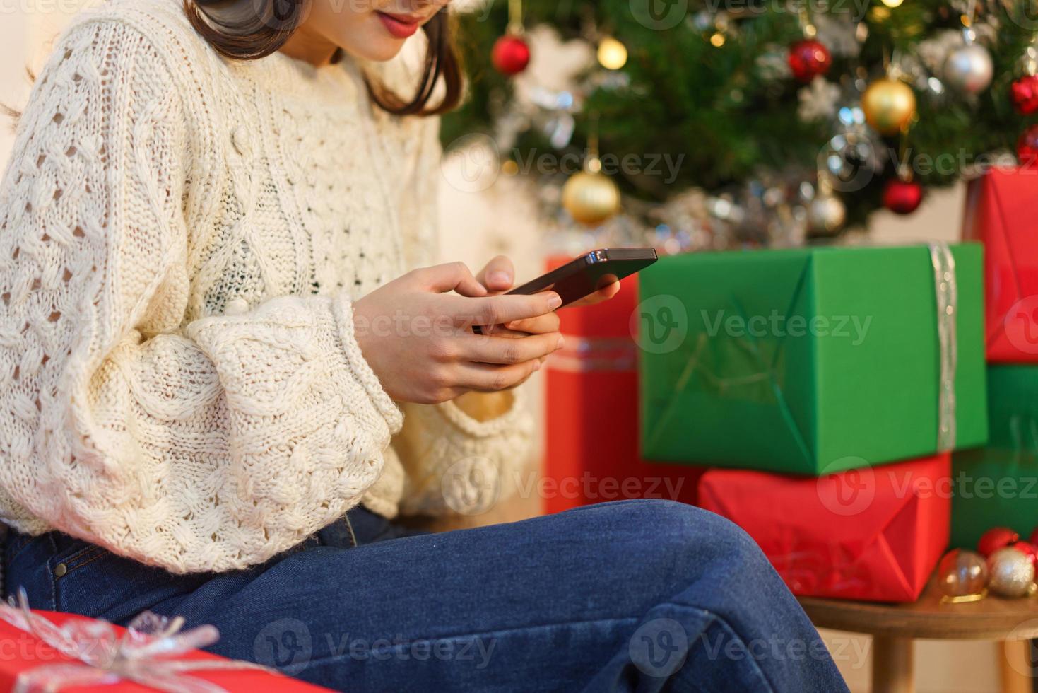 Natale concetto, asiatico donna si siede nel Natale decorato vivente camera e chat su smartphone foto
