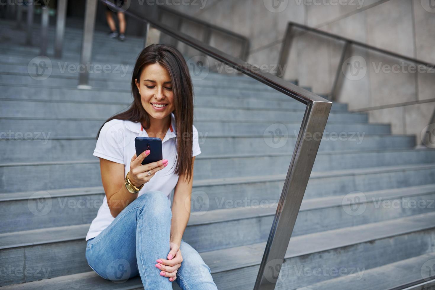 seduta su il le scale con smartphone nel mani. bella giovane donna avere fine settimana nel il città a giorno foto