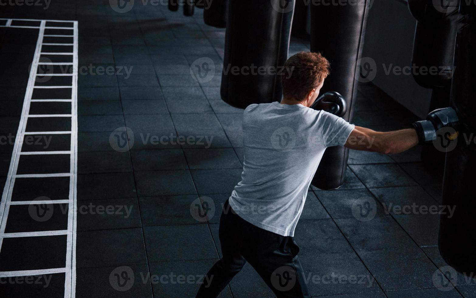 giovane uomo nel bianca camicia e boxe protettivo guanti fare esercizi nel Palestra con spingendo Borsa foto