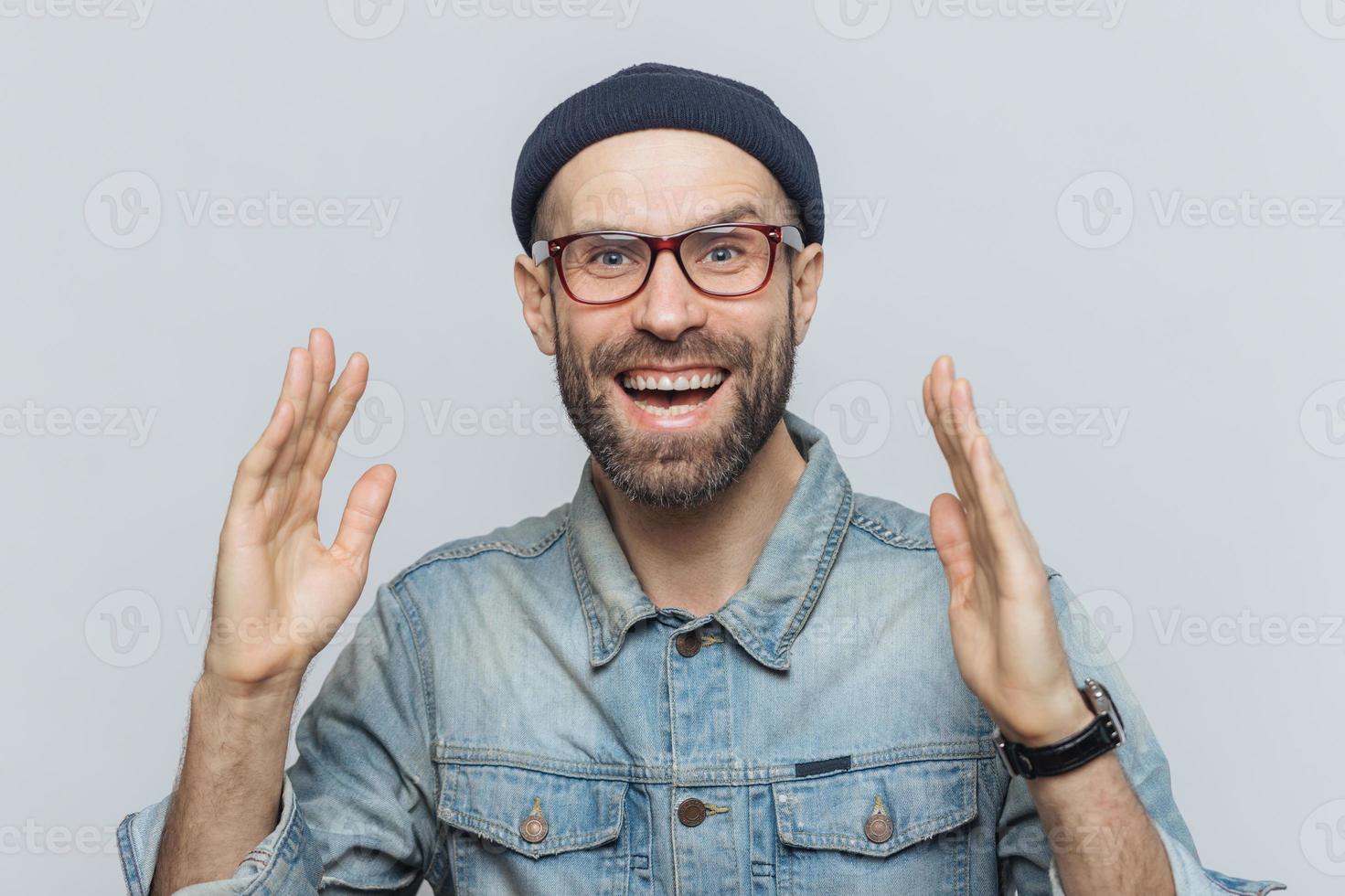 bell'uomo felice alza le mani con eccitazione, ha un'espressione felicissima, guarda con una faccia felice, ha folta barba e baffi. uomo felice in occhiali pone su sfondo bianco, si diverte foto