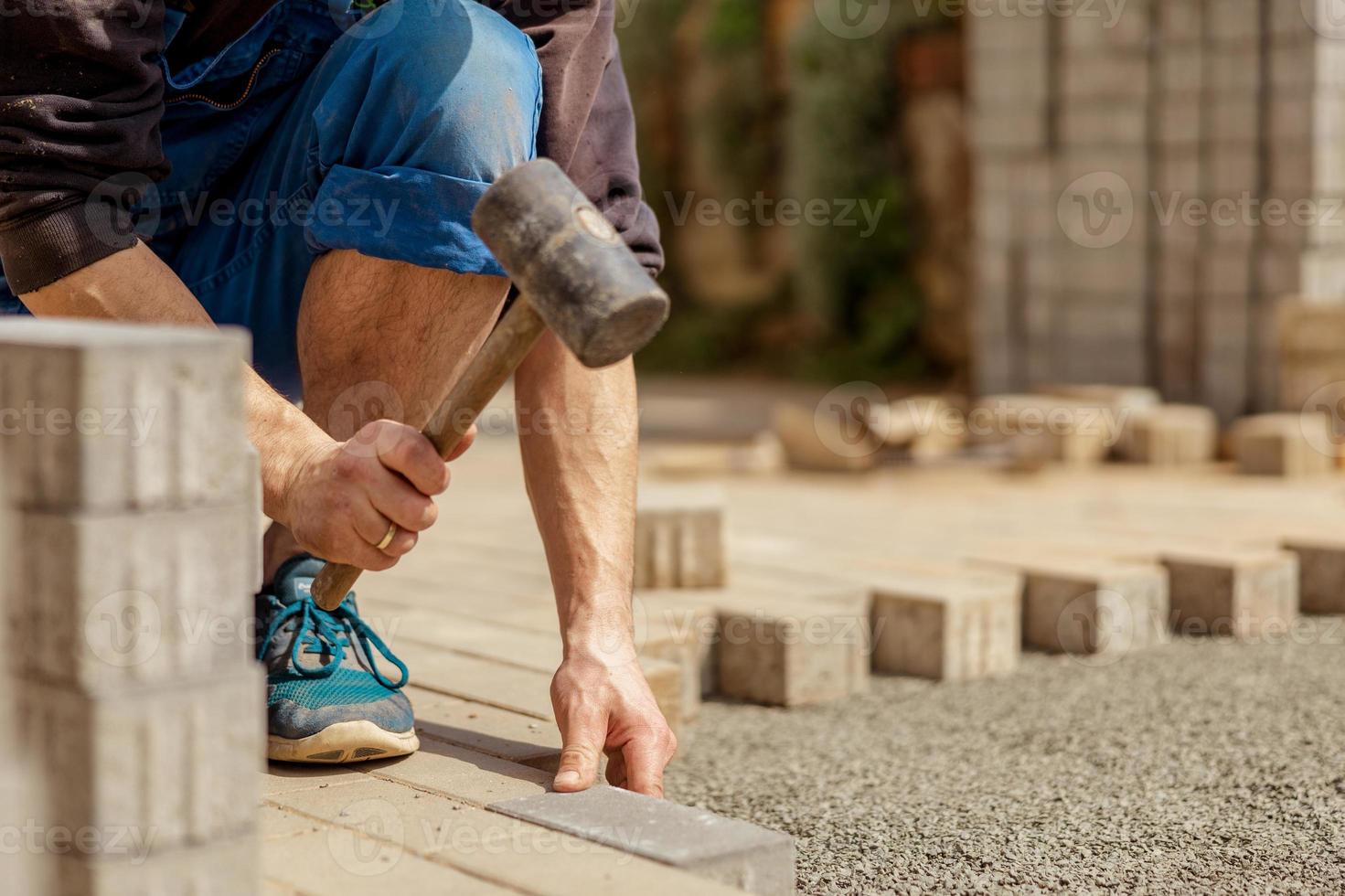 giovane uomo posa grigio calcestruzzo pavimentazione lastre nel Casa cortile su ghiaia fondazione base. maestro stabilisce pavimentazione pietre. giardino mattone sentiero pavimentazione di professionale finitrice lavoratore. riparazione marciapiede. foto