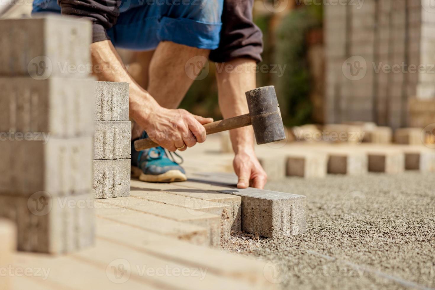 giovane uomo posa grigio calcestruzzo pavimentazione lastre nel Casa cortile su ghiaia fondazione base. maestro stabilisce pavimentazione pietre. giardino mattone sentiero pavimentazione di professionale finitrice lavoratore. riparazione marciapiede. foto