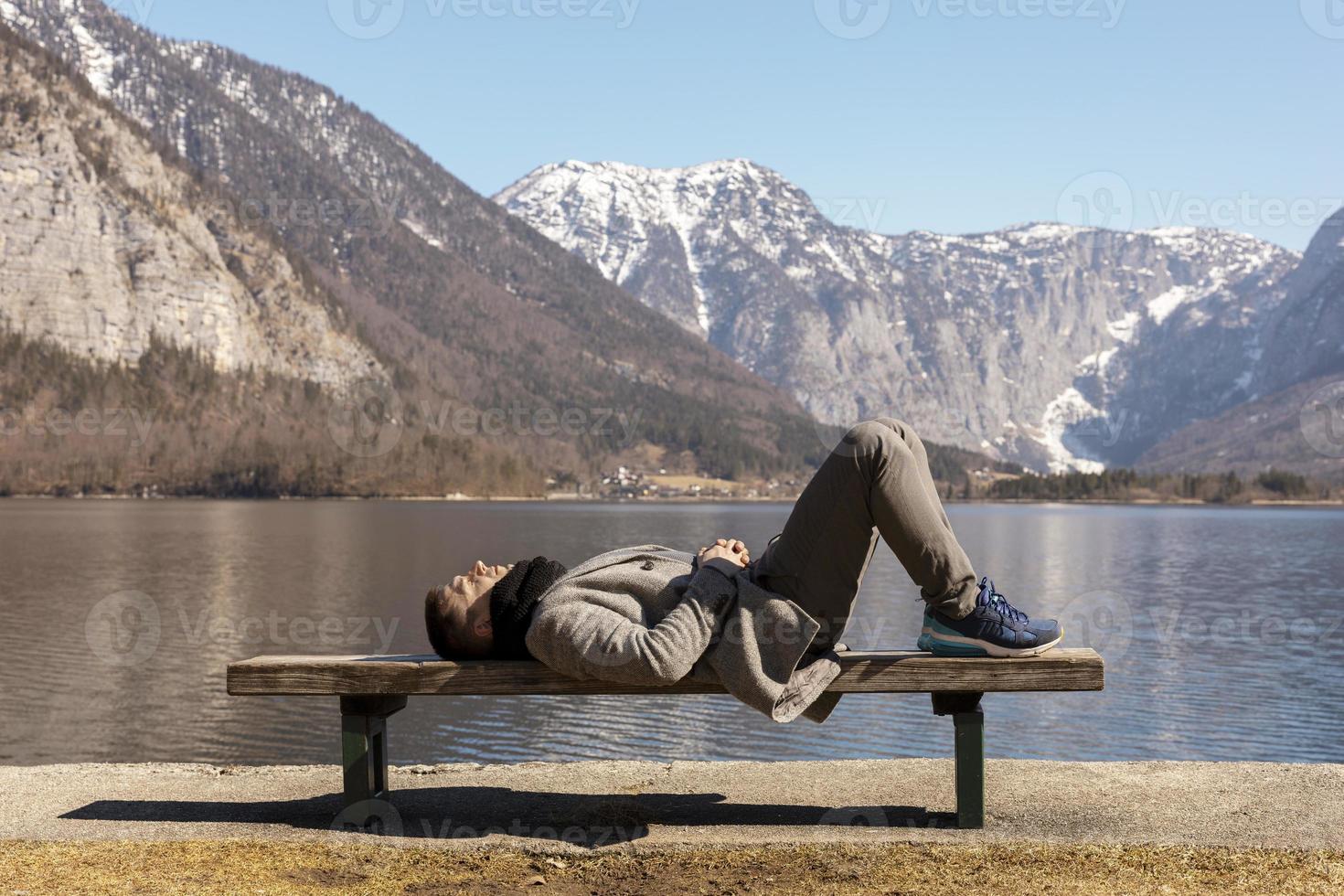 giovane uomo dire bugie all'aperto su panchina e godendo montagne, neve, bene tempo atmosferico, blu cielo, sole. bellissimo paesaggio. tempo con te stesso, sognando, rilassamento, mentale Salute. turismo, vacanza, viaggio. foto