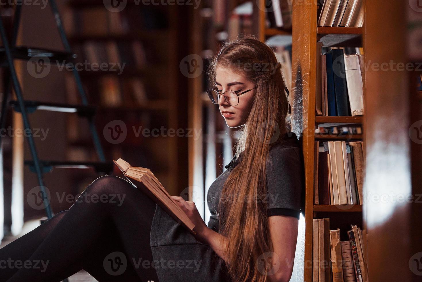 si siede su il pavimento. femmina alunno è nel biblioteca quello pieno di libri. concezione di formazione scolastica foto