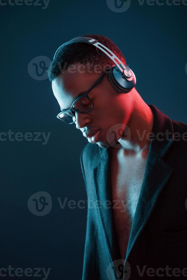 godendo ascoltando musica nel cuffia. nel occhiali. futuristico neon illuminazione. giovane africano americano uomo nel il studio foto