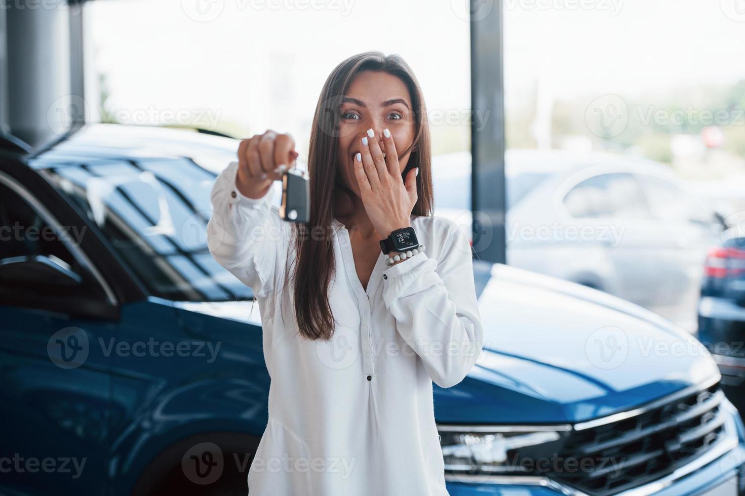 eccitato con sua nuovo macchina. detiene chiavi nel mano. giovane donna nel bianca ufficiale Abiti sta nel davanti di blu automobile in casa foto