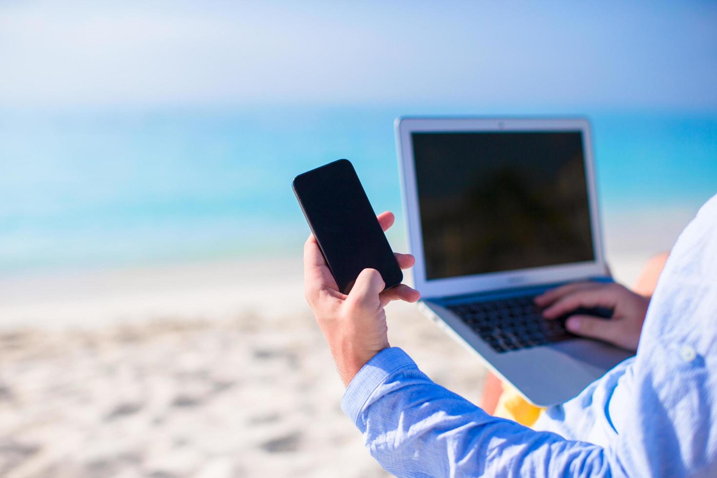 primo piano di una persona che utilizza un telefono e un laptop su una spiaggia foto
