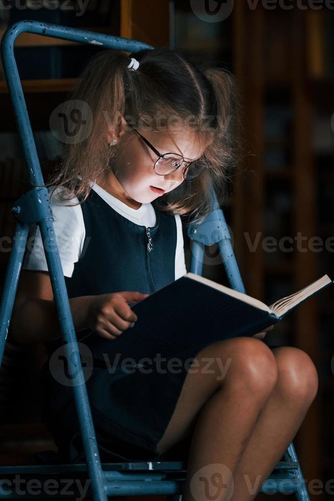 concentrazione su lettura. scuola ragazza su il scala nel biblioteca pieno di libri. formazione scolastica concezione foto