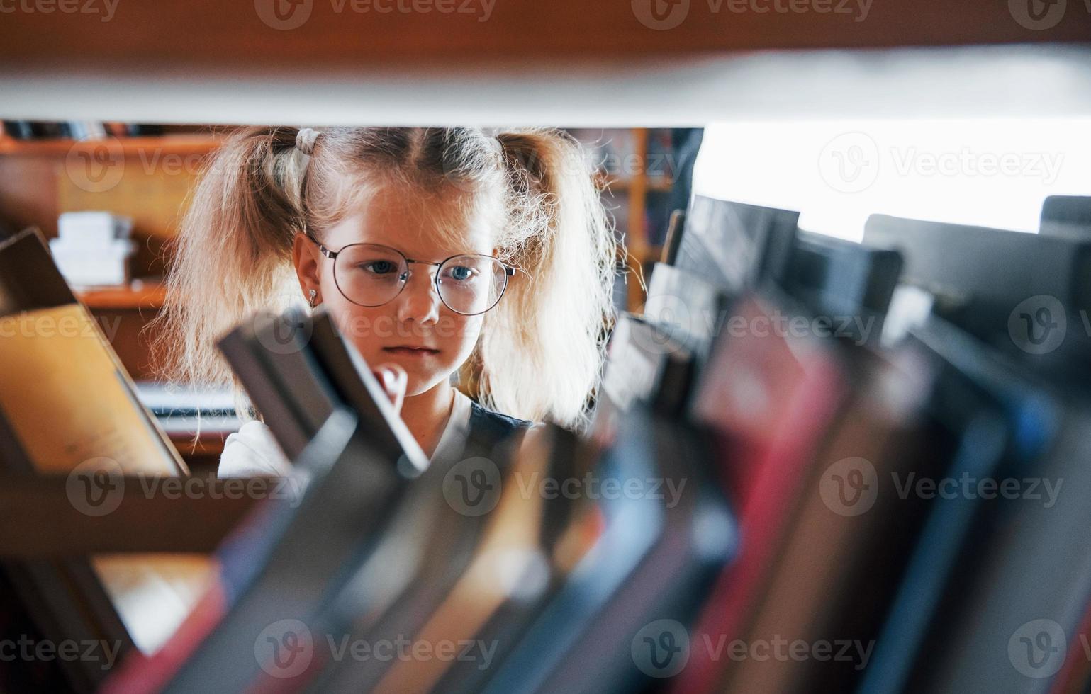 poco ragazza nel bicchieri ricerca per un' libro nel il biblioteca. concezione di formazione scolastica foto