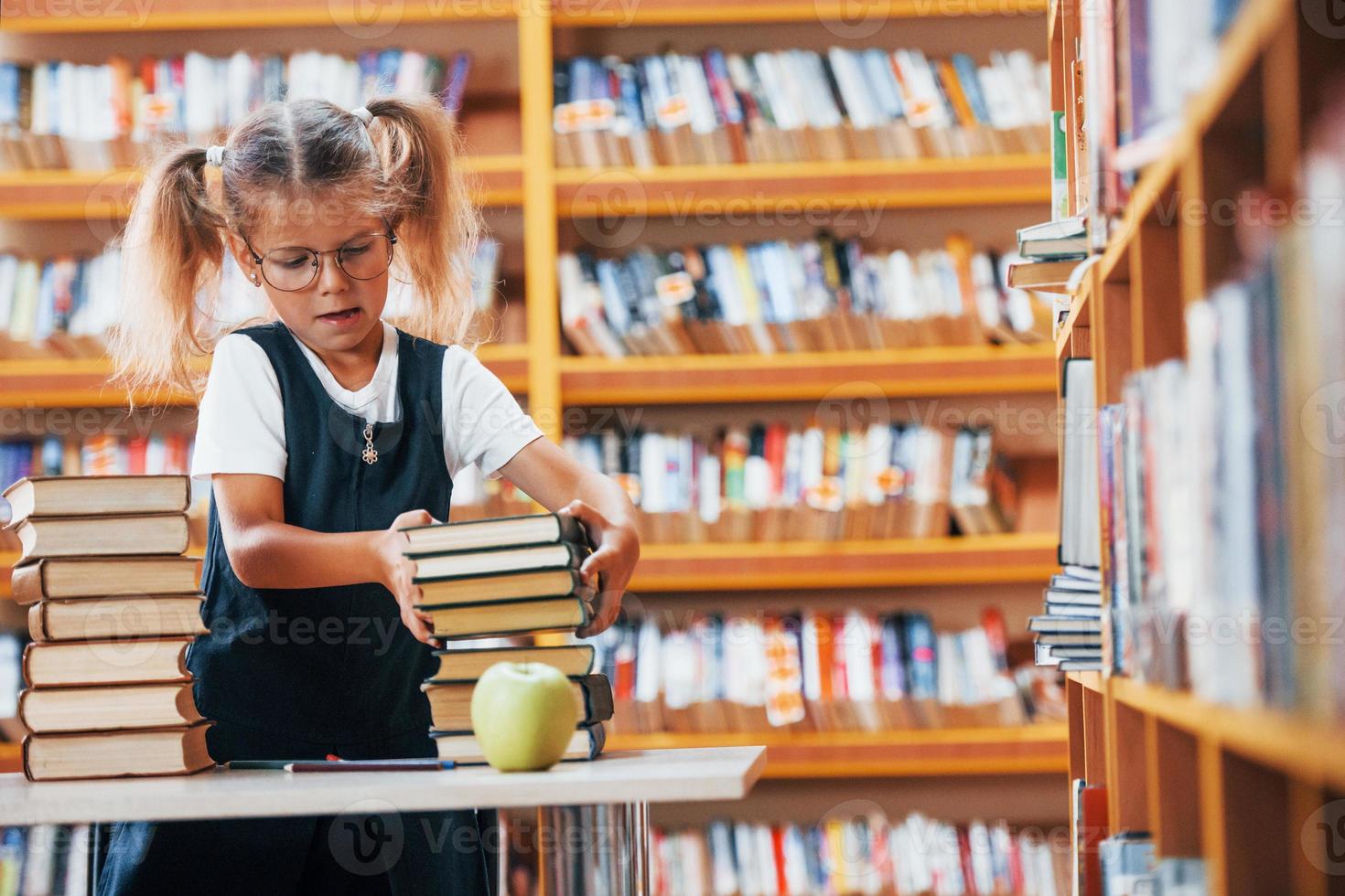 carino poco ragazza con trecce è nel il biblioteca. Mela su il libri foto