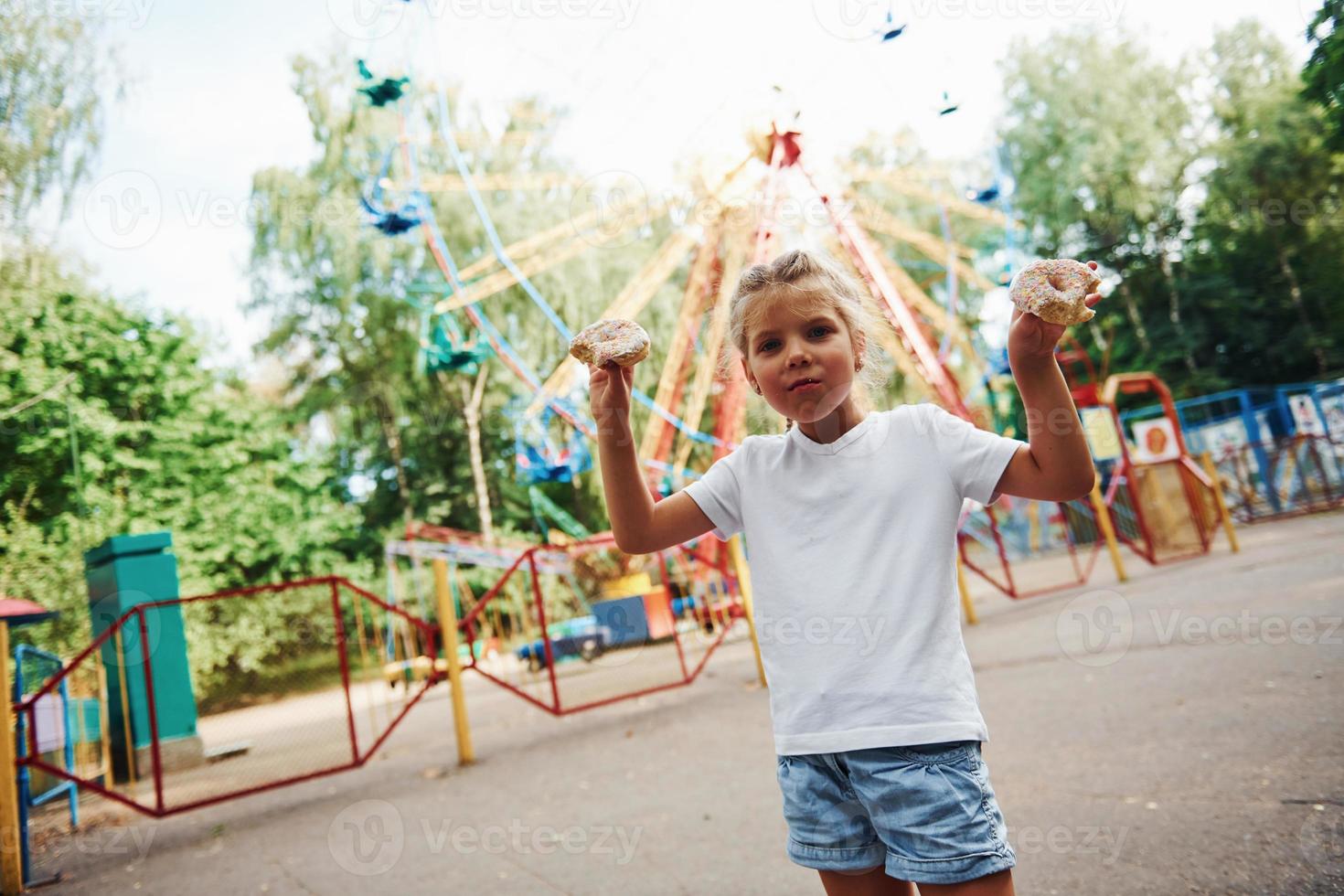 carino poco ragazza mangia ciambelle nel il parco a giorno vicino attrazioni foto