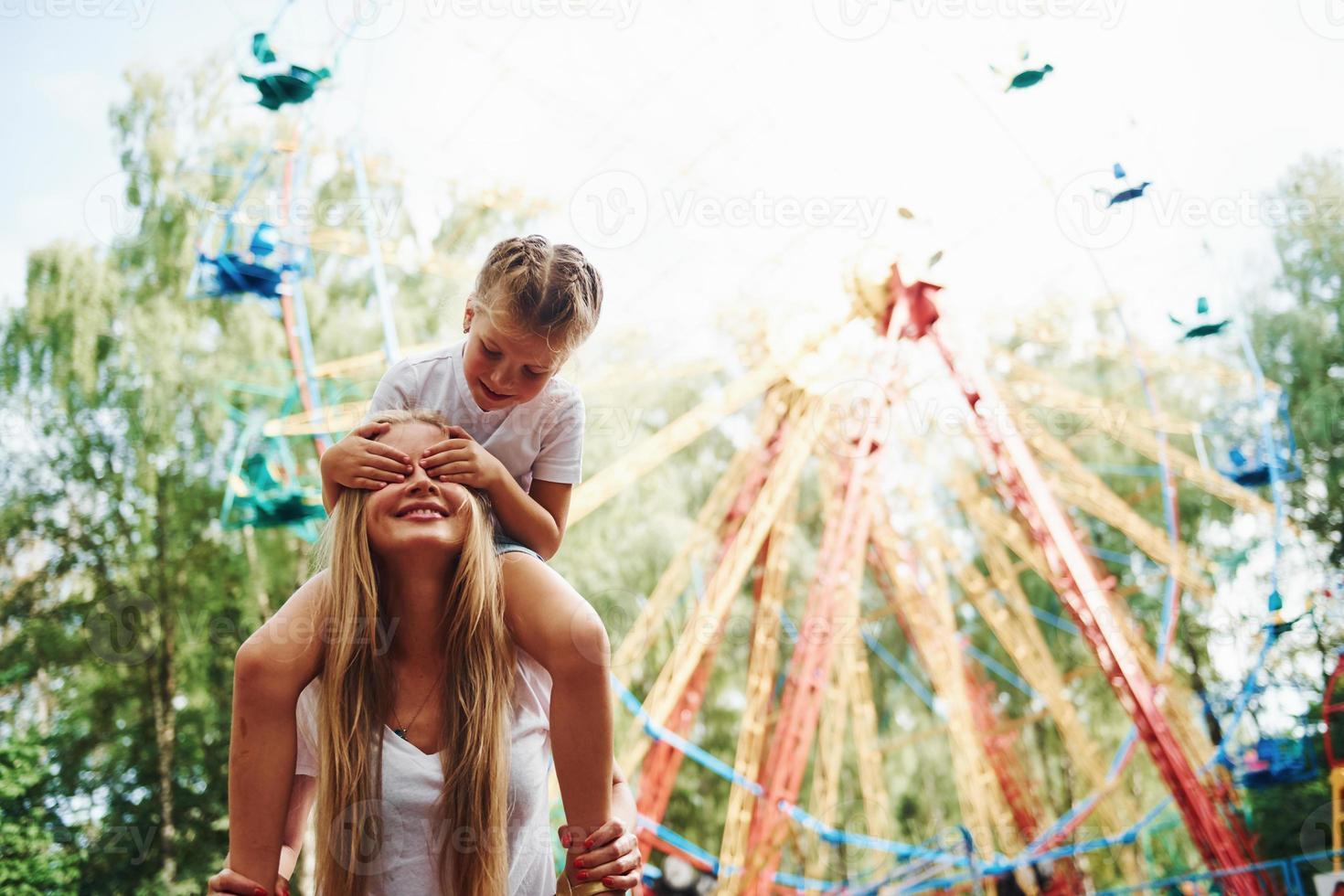 figlia si siede su il le spalle. allegro poco ragazza sua madre avere un' bene tempo nel il parco insieme vicino attrazioni foto