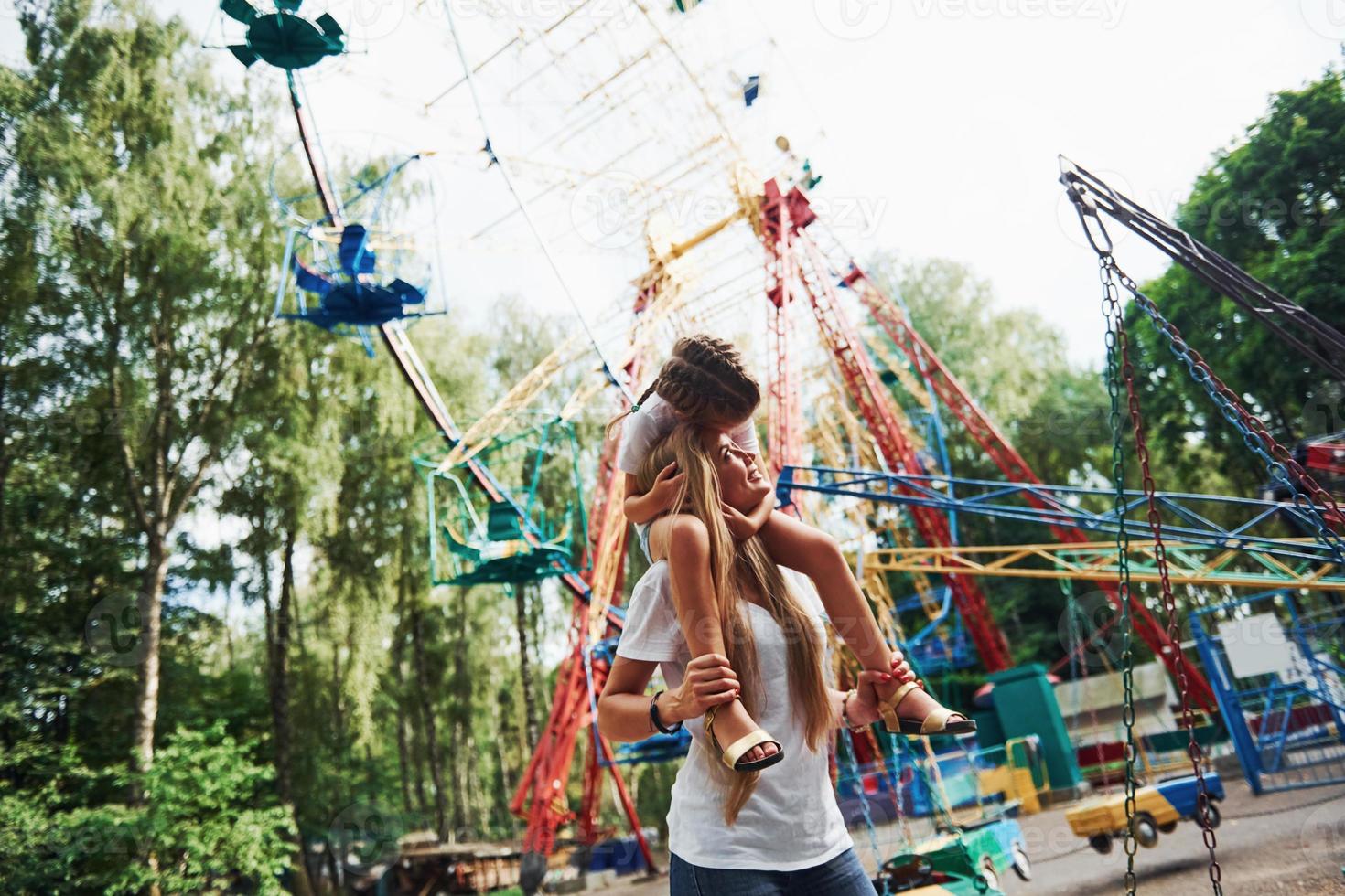 a piedi con figlia su il le spalle. allegro poco ragazza sua madre avere un' bene tempo nel il parco insieme vicino attrazioni foto