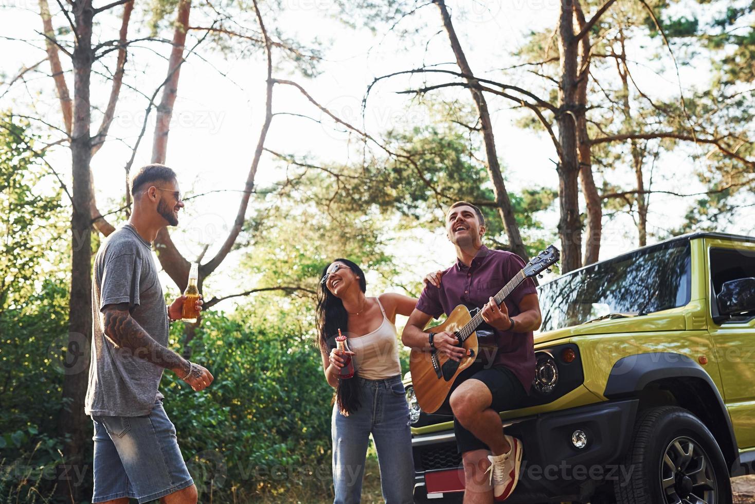 cantando canzoni insieme. amici avere simpatico fine settimana all'aperto vicino il loro verde auto con acustico chitarra foto