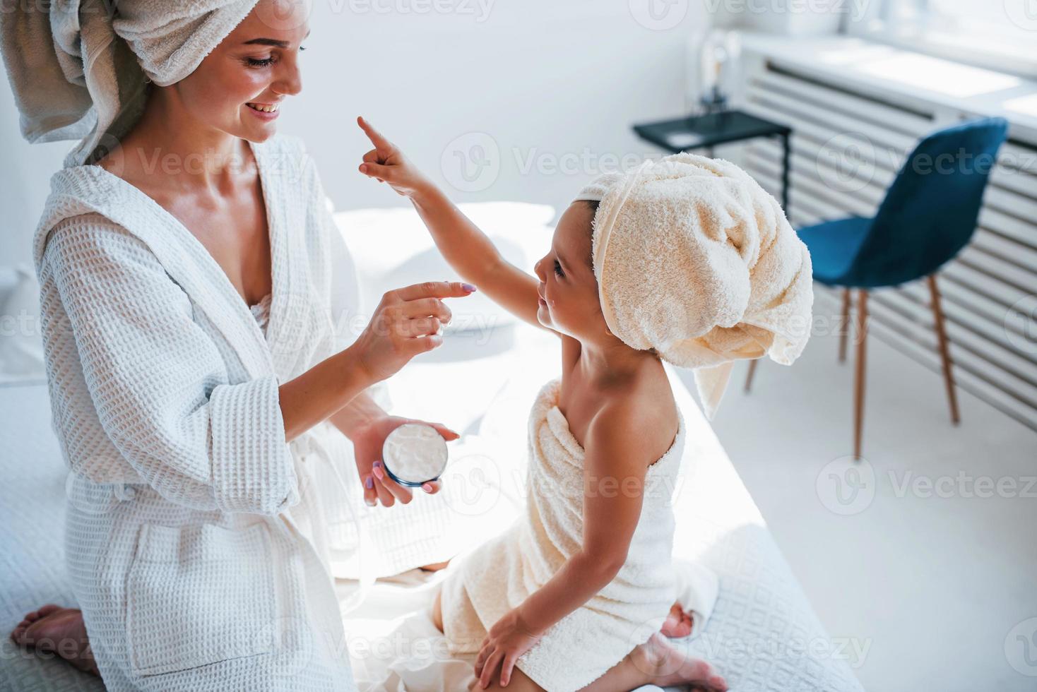 utilizzando crema per chiaro pelle. giovane madre con sua figlia avere bellezza giorno in casa nel bianca camera foto