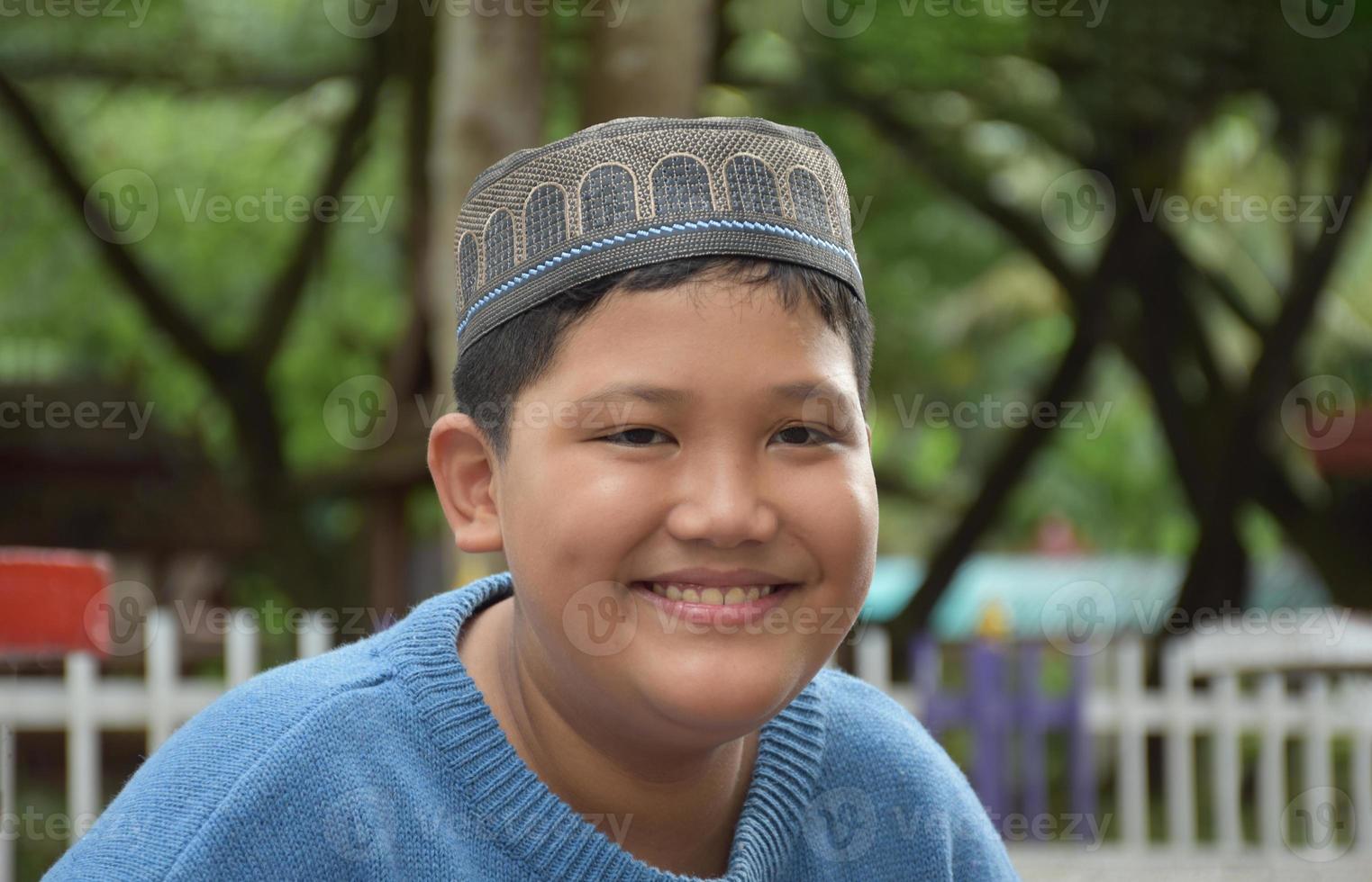 ritratto asiatico musulmano o islamico ragazzo seduta nel il scuola parco e sorridente felicemente, morbido e selettivo messa a fuoco. foto