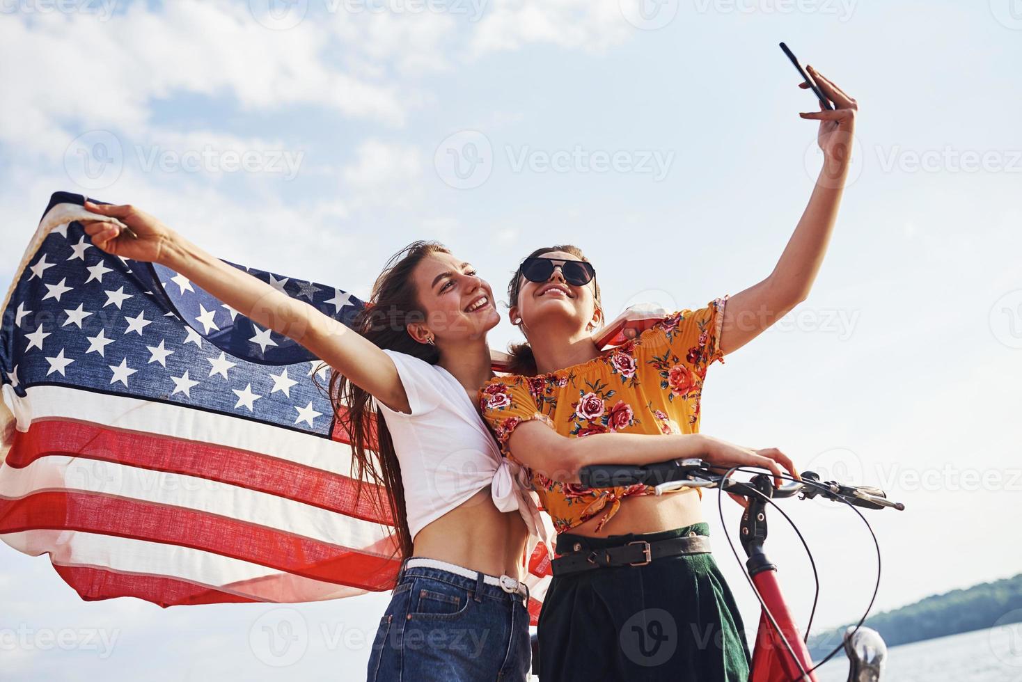 Due patriottico allegro donne con bicicletta e Stati Uniti d'America bandiera nel mani fa autoscatto foto