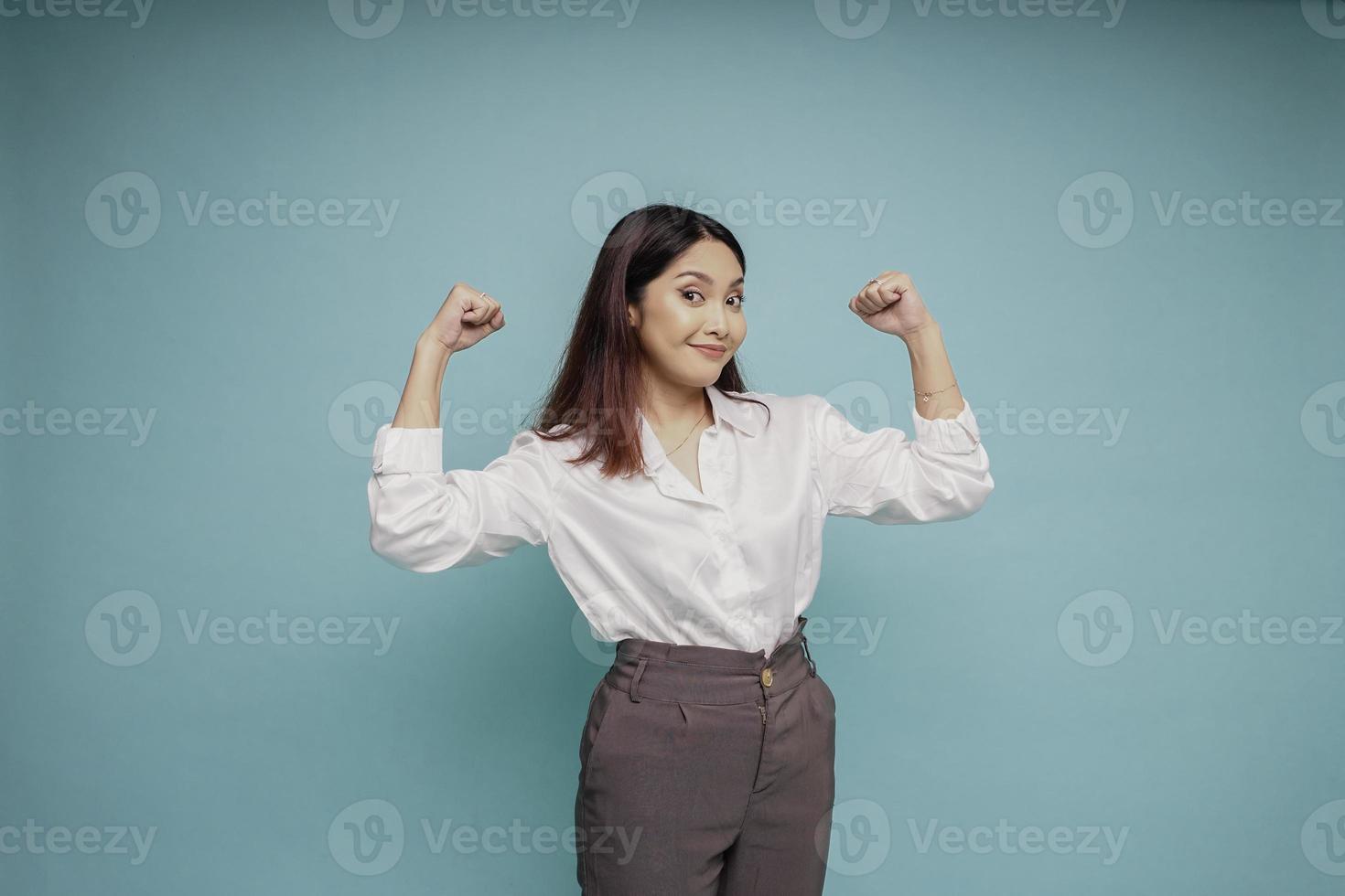 eccitato asiatico donna indossare un' bianca camicia mostrando forte gesto di sollevamento sua braccia e muscoli sorridente con orgoglio foto