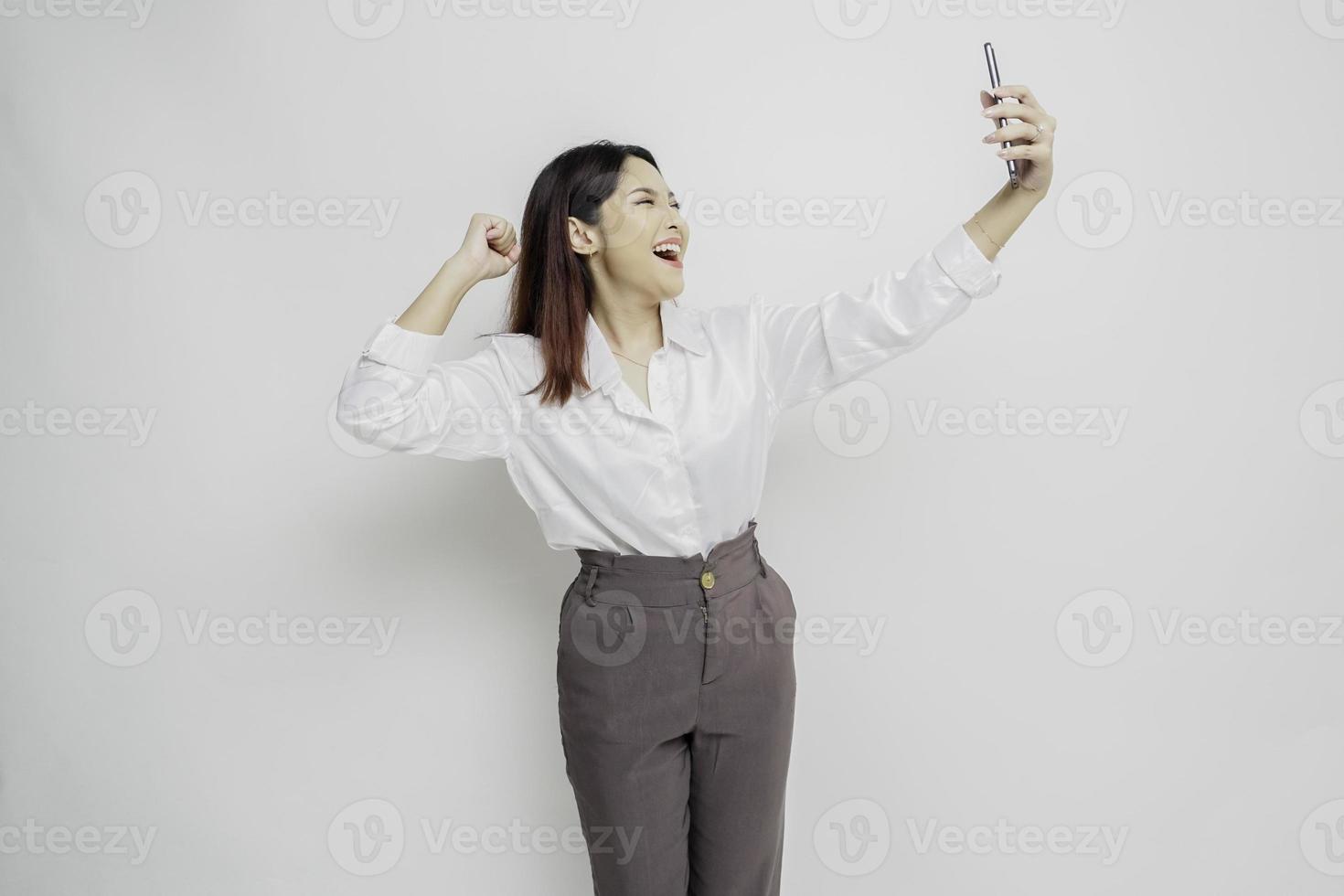 un' giovane asiatico donna con un' contento riuscito espressione indossare bianca camicia e Tenere smartphone isolato di bianca sfondo foto